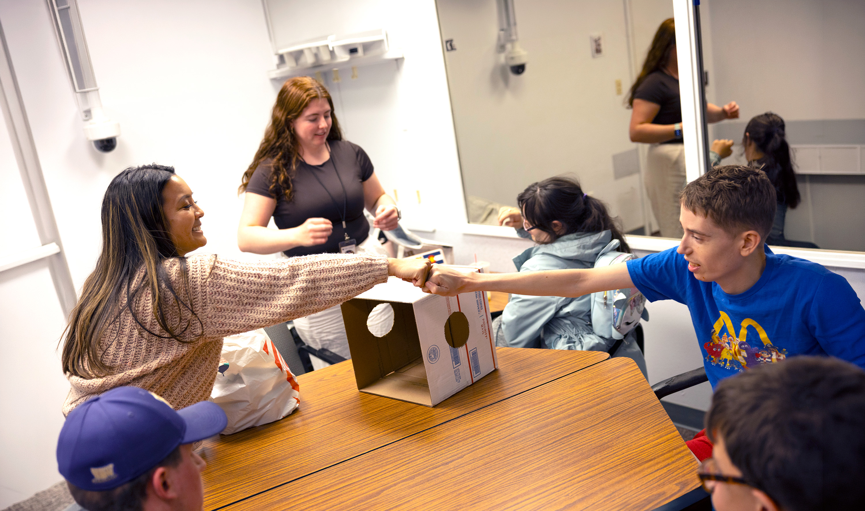 People interact around a table, with two of them reaching across for a fist bump