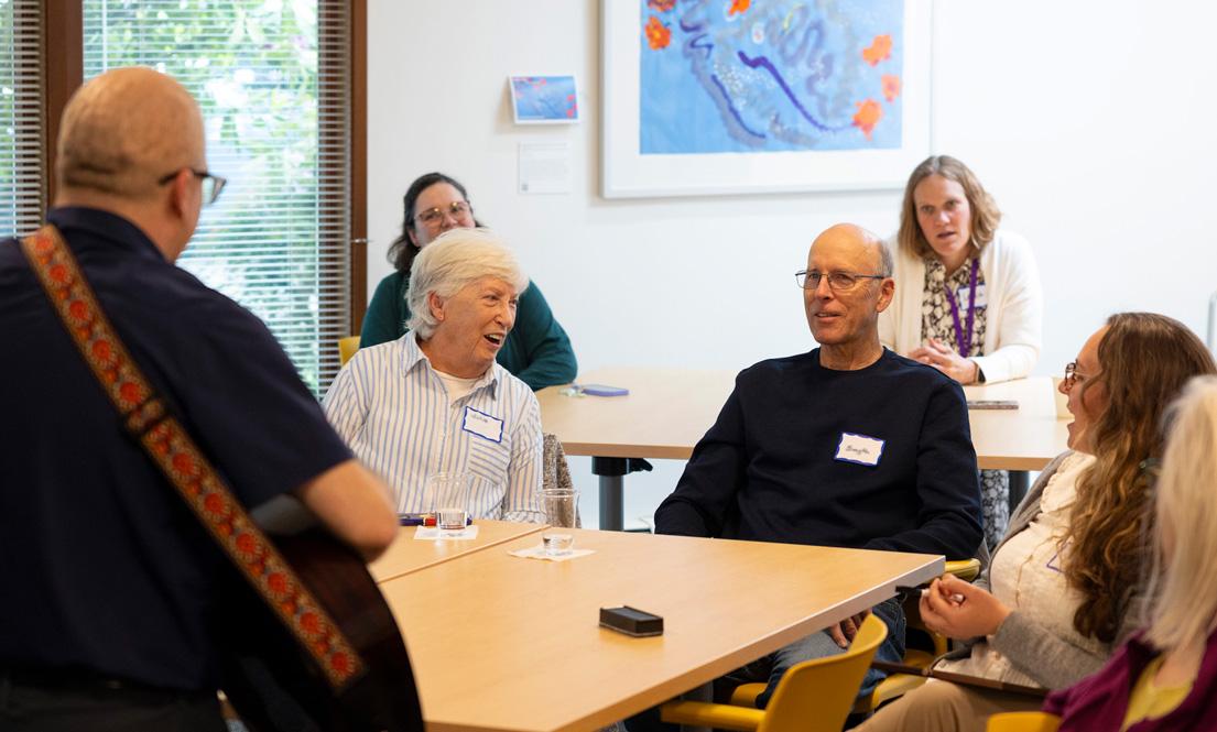A small group sits around a table, listening to a person playing guitar, during a Memory Hub event