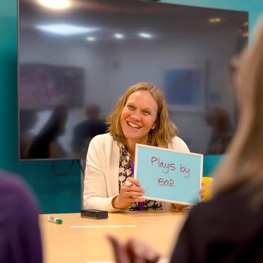 A woman smiles while holding up a whiteboard that says “Plays by ear” during a group activity.